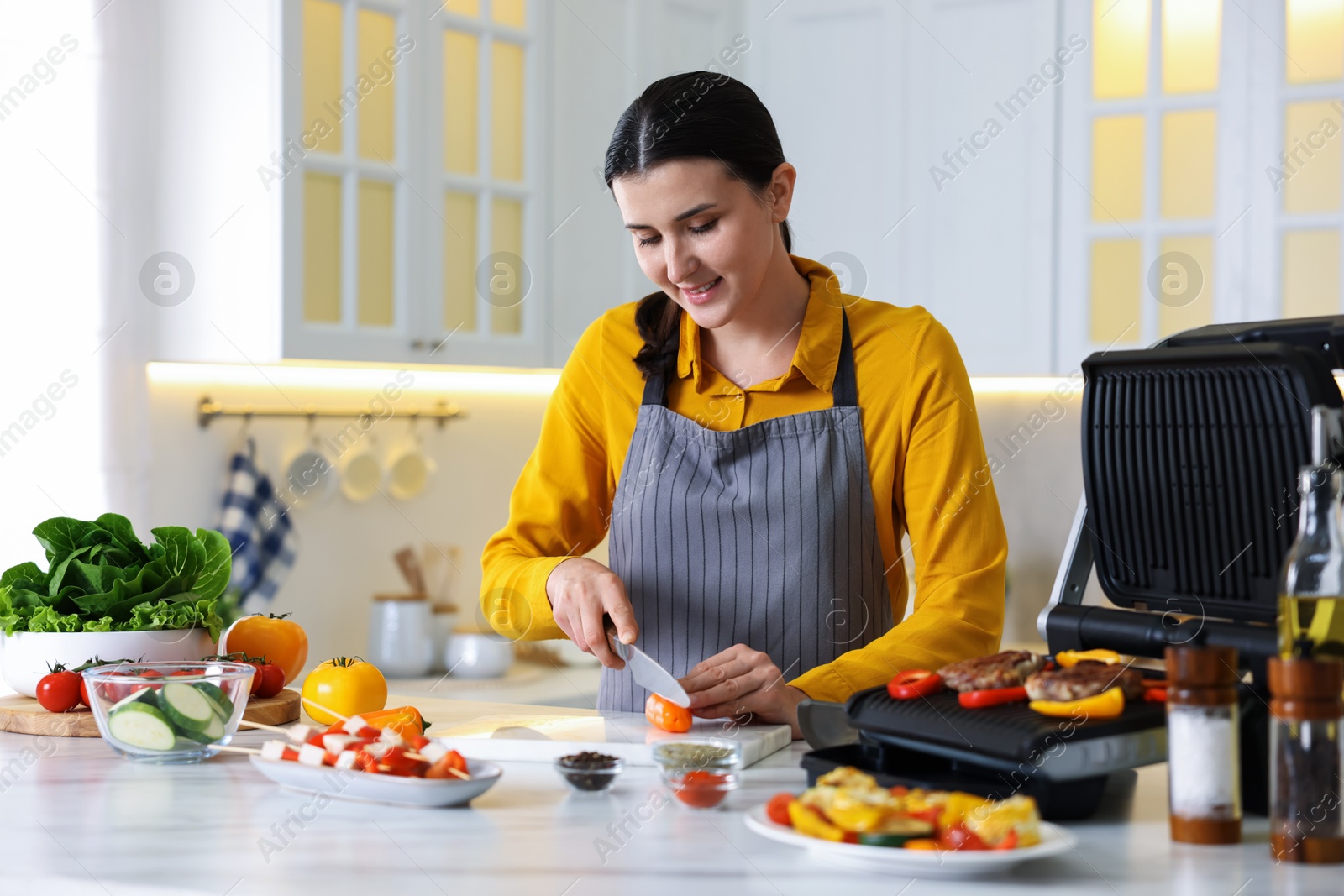 Photo of Woman cutting pepper while cooking meat and vegetables on electric grill in kitchen
