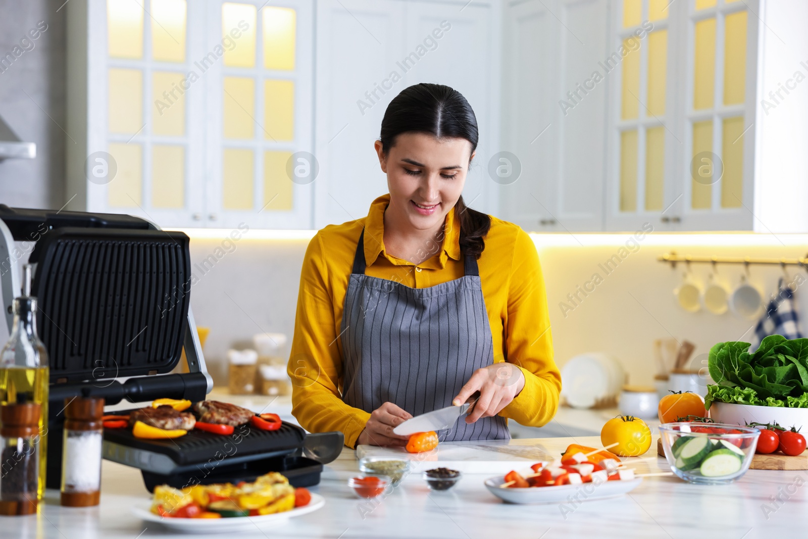 Photo of Woman cutting pepper while cooking meat and vegetables on electric grill in kitchen