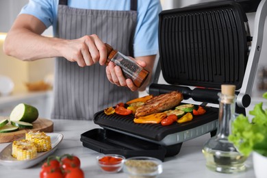 Photo of Man adding pepper to vegetables and meat cooking on electric grill in kitchen, closeup