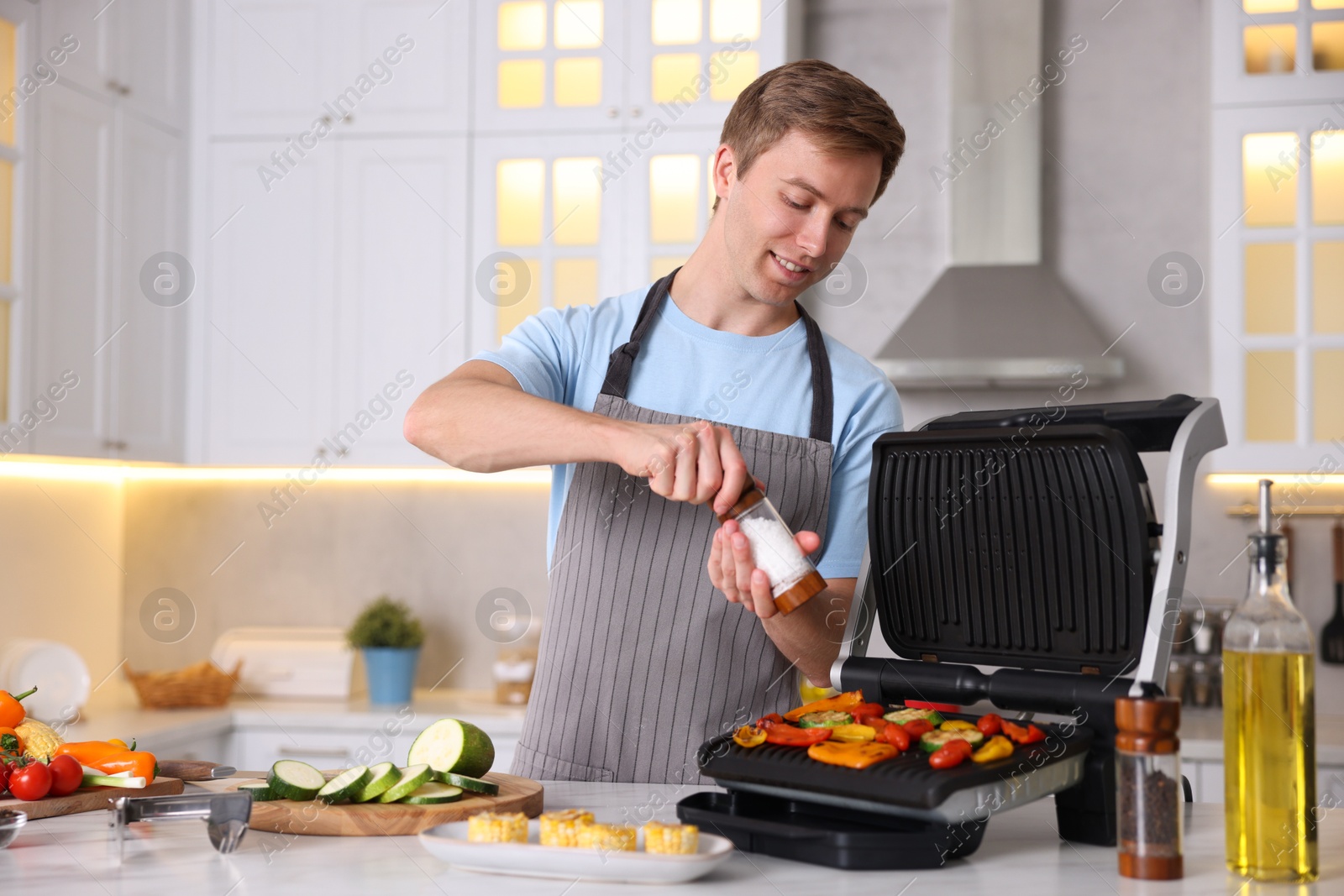 Photo of Man adding salt to vegetables cooking on electric grill in kitchen