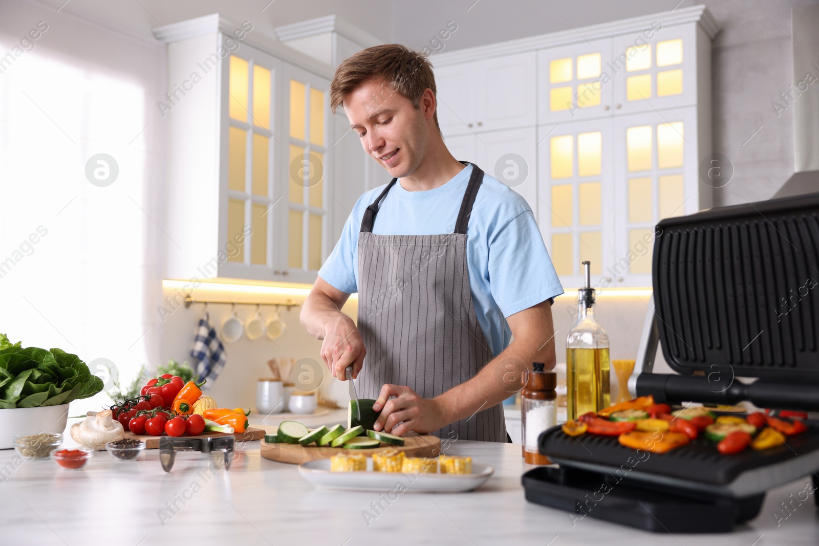 Photo of Man cutting zucchini while cooking vegetables on electric grill in kitchen
