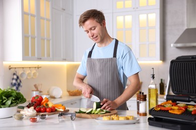 Photo of Man cutting zucchini while cooking vegetables on electric grill in kitchen