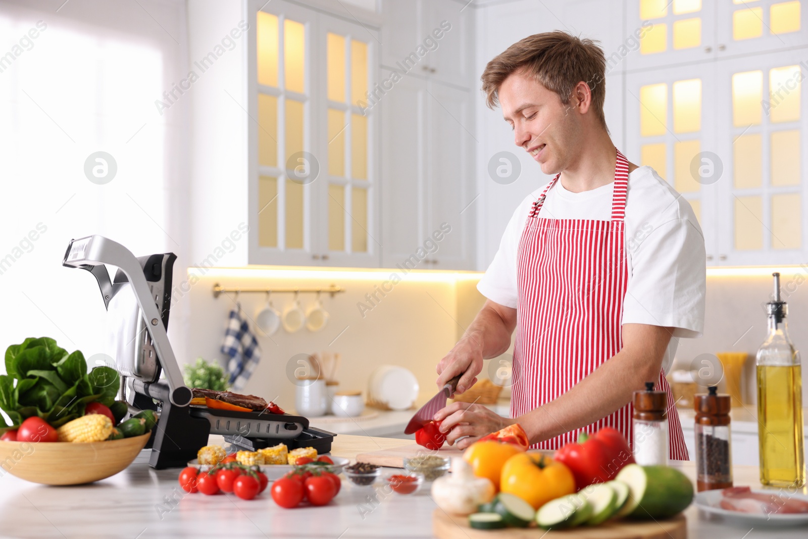 Photo of Man cutting pepper while cooking vegetables on electric grill in kitchen