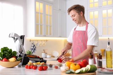 Photo of Man cutting pepper while cooking vegetables on electric grill in kitchen