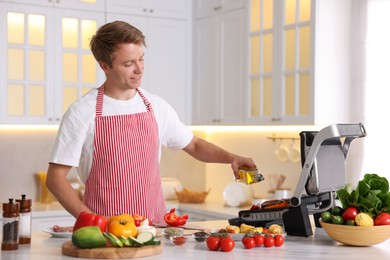 Photo of Man pouring oil onto vegetables cooking on electric grill in kitchen