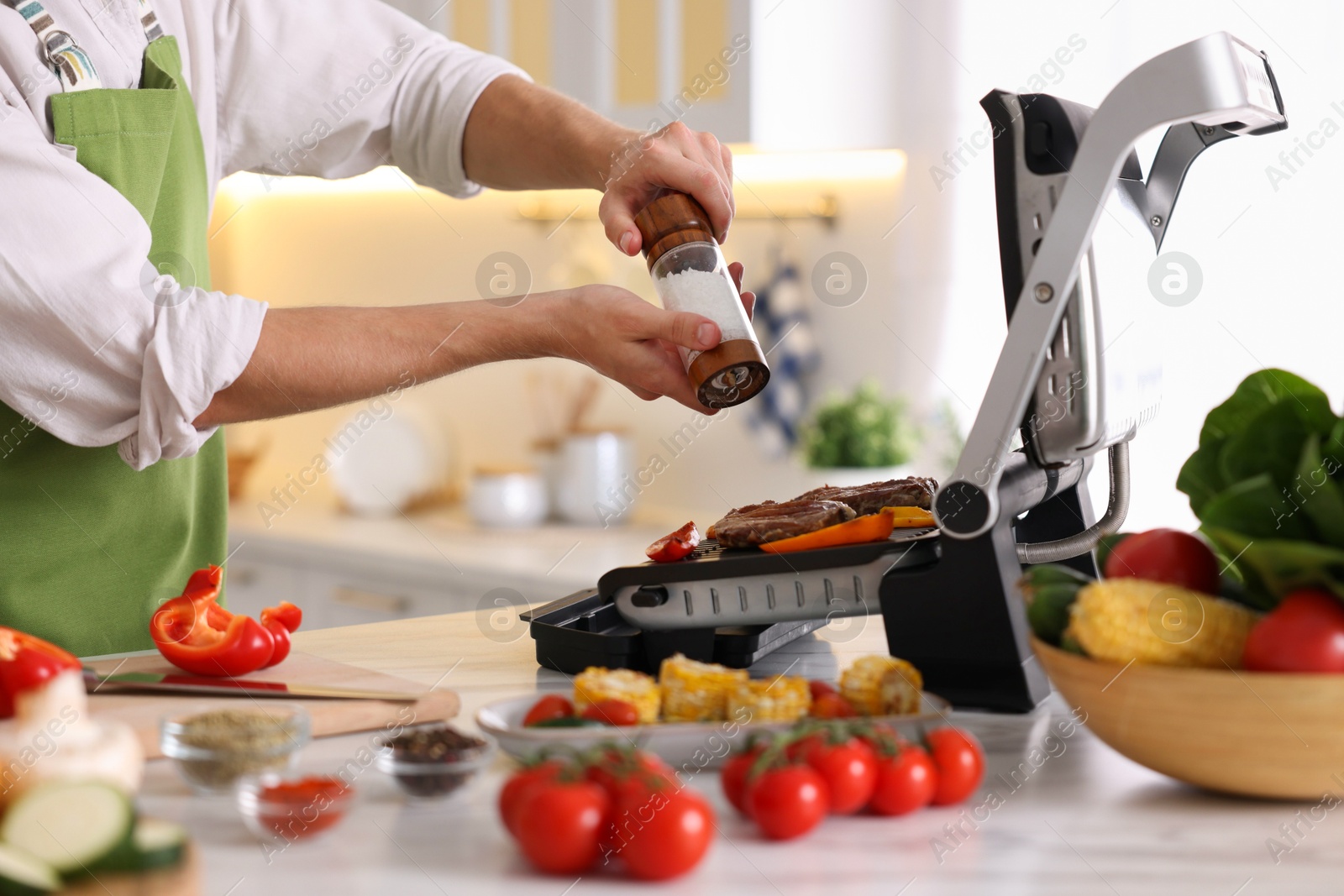Photo of Man adding salt to vegetables and meat cooking on electric grill in kitchen, closeup