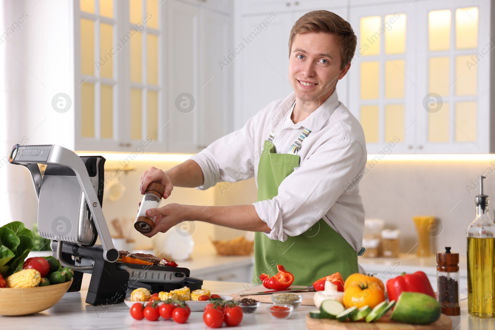Photo of Man adding salt to vegetables and meat cooking on electric grill in kitchen