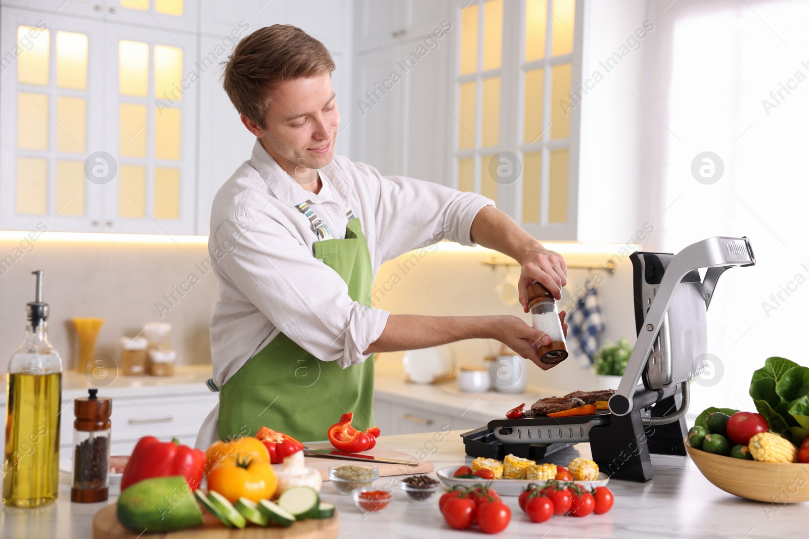 Photo of Man adding salt to vegetables and meat cooking on electric grill in kitchen