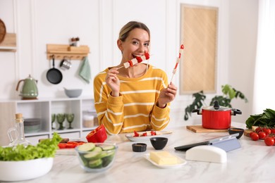 Photo of Smiling woman eating tasty skewers after cooking at table in kitchen