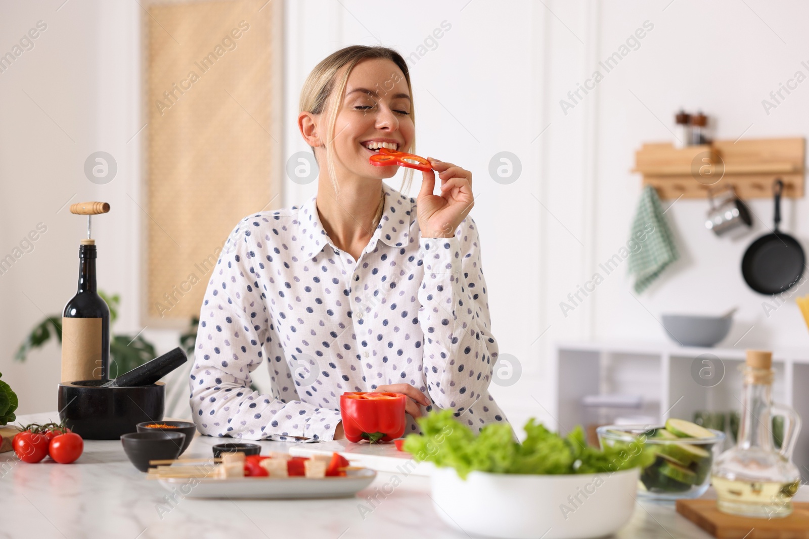 Photo of Smiling woman eating bell pepper while cooking at table in kitchen