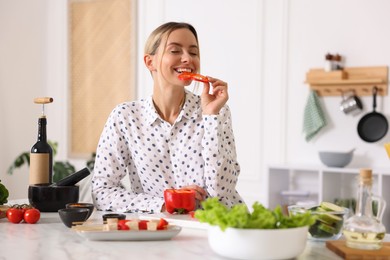 Photo of Smiling woman eating bell pepper while cooking at table in kitchen