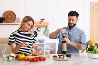 Photo of Smiling man opening bottle of wine while cooking with his wife at table in kitchen