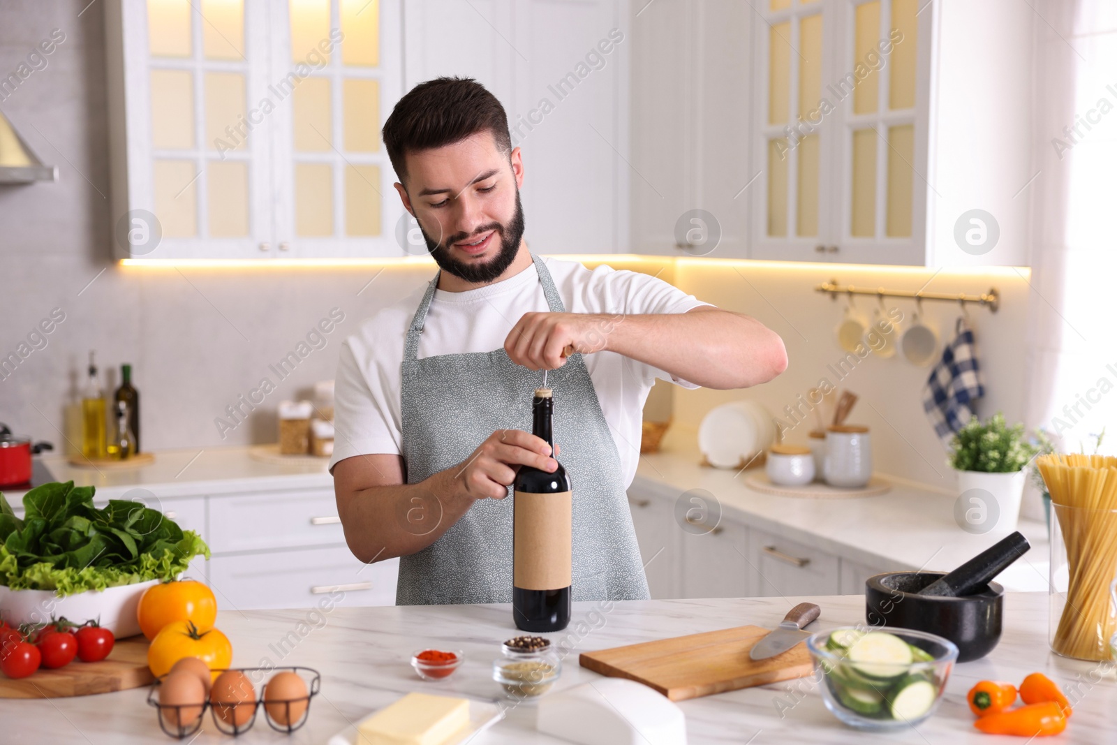 Photo of Smiling man opening bottle of wine while cooking at table in kitchen