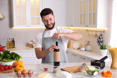 Photo of Smiling man opening bottle of wine while cooking at table in kitchen