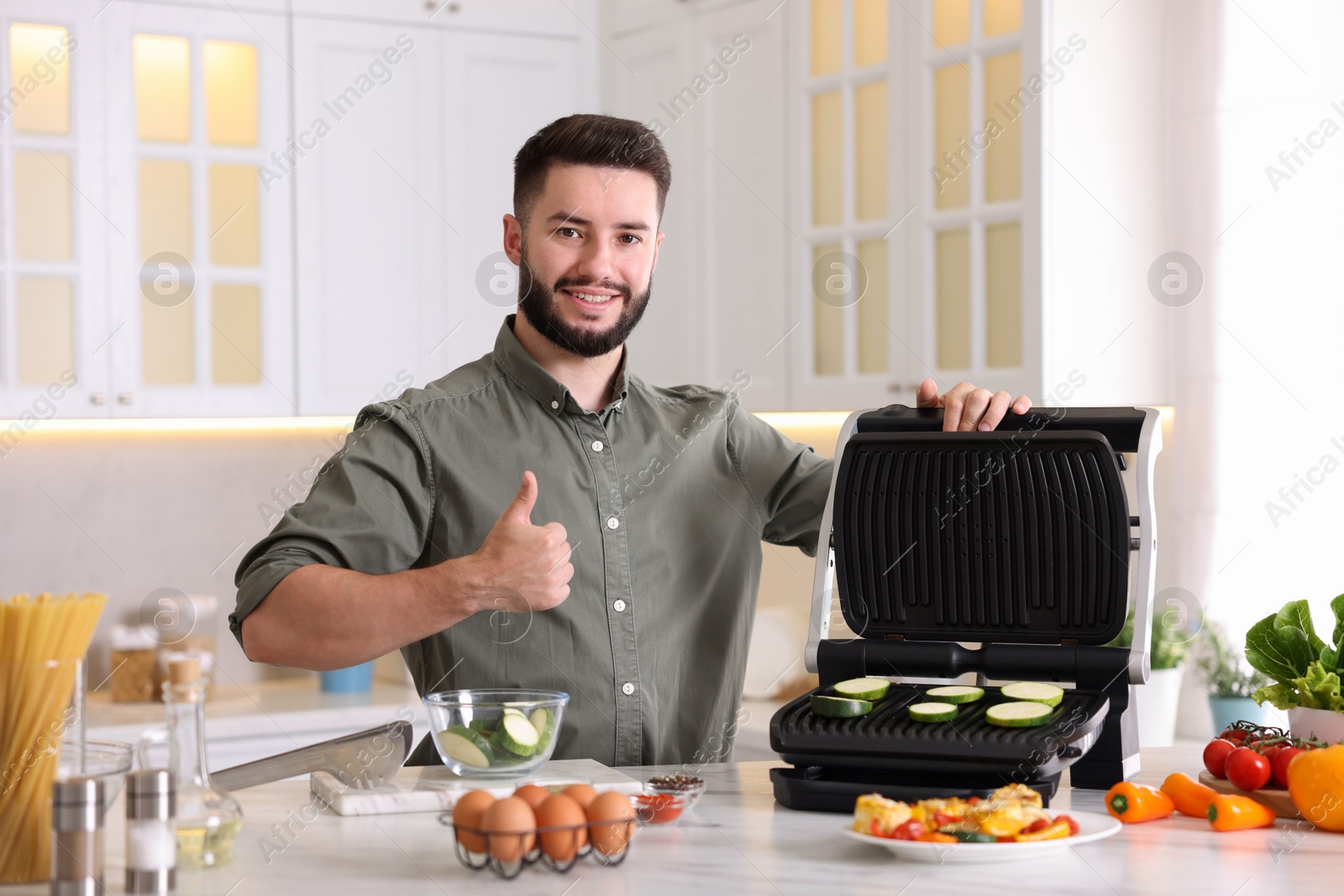 Photo of Smiling man cooking zucchini on electric grill and showing thumbs up at table in kitchen