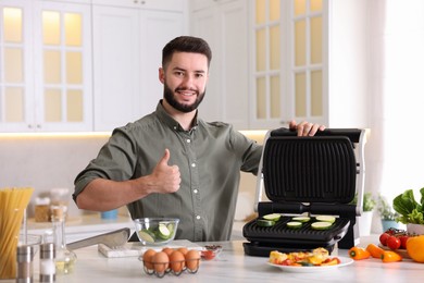 Photo of Smiling man cooking zucchini on electric grill and showing thumbs up at table in kitchen