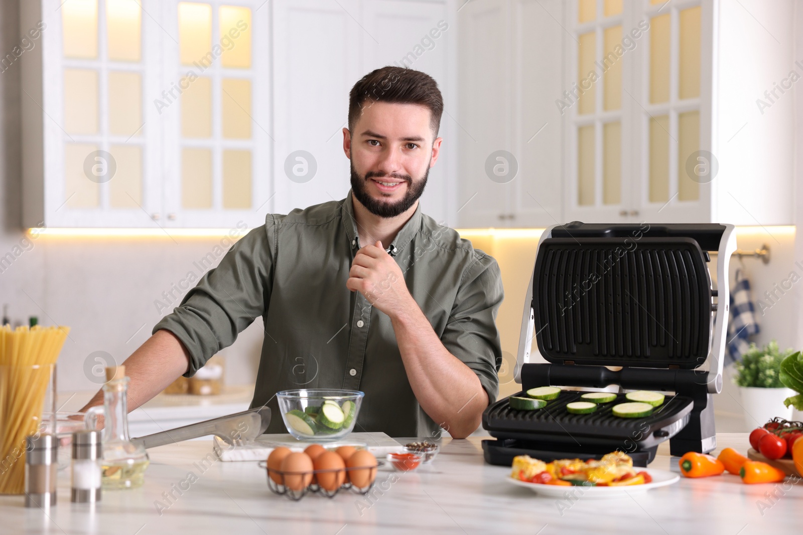 Photo of Smiling man cooking zucchini on electric grill at table in kitchen