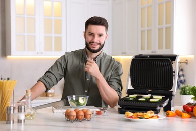 Photo of Smiling man cooking zucchini on electric grill at table in kitchen
