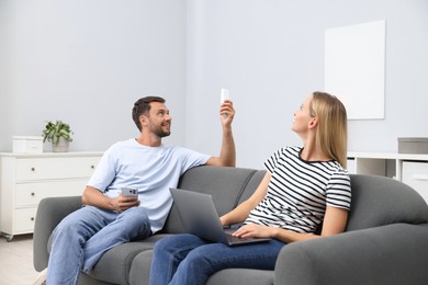Photo of Couple with laptop and smartphone using remote to control air conditioner on sofa at home