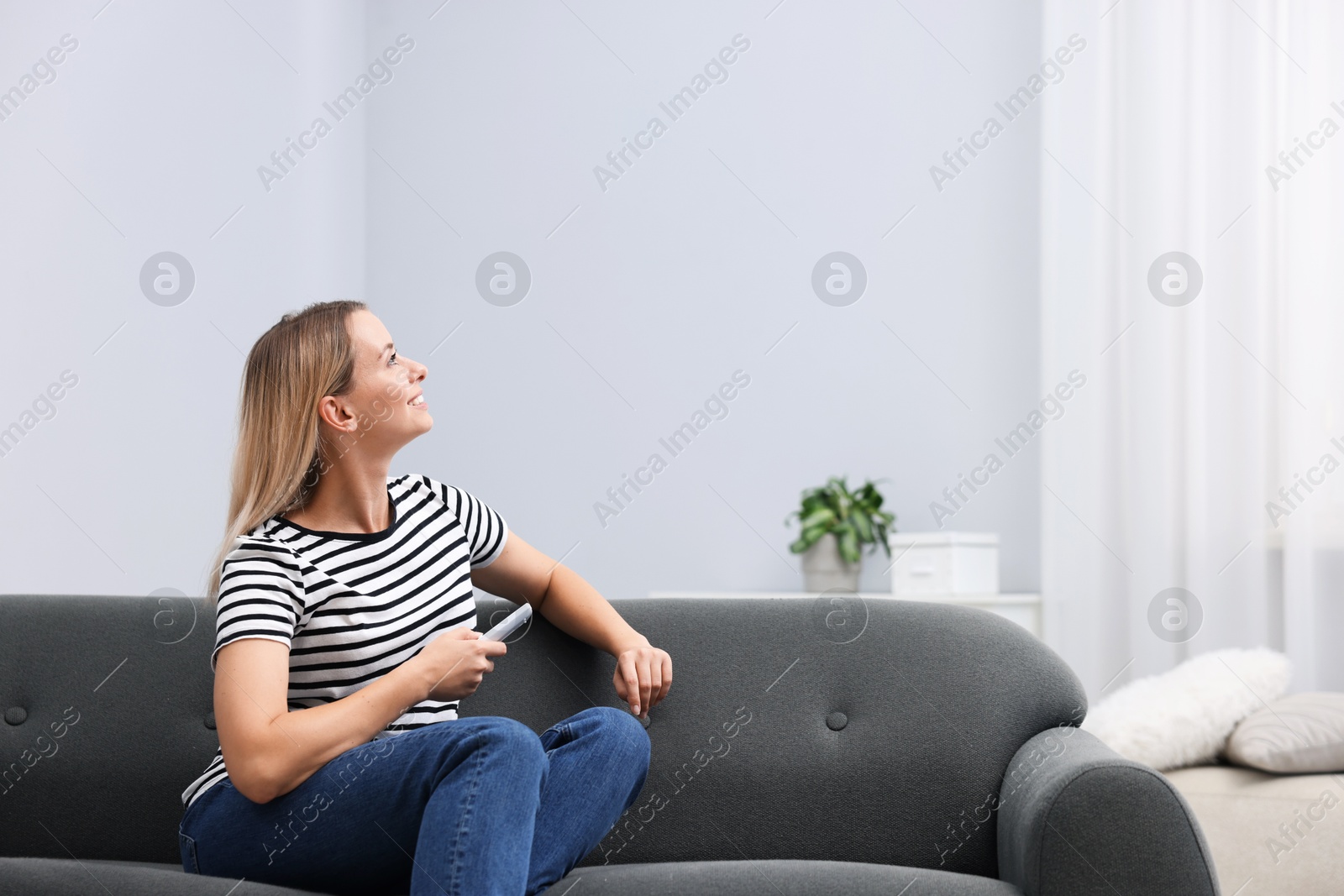 Photo of Woman using remote to control air conditioner on sofa at home. Space for text