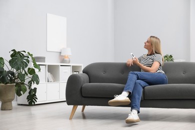 Photo of Woman using remote to control air conditioner on sofa at home. Space for text