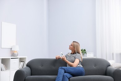 Photo of Woman using remote to control air conditioner on sofa at home. Space for text