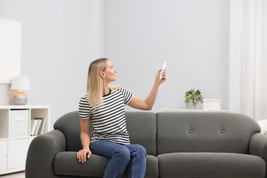 Photo of Woman using remote to control air conditioner on sofa at home