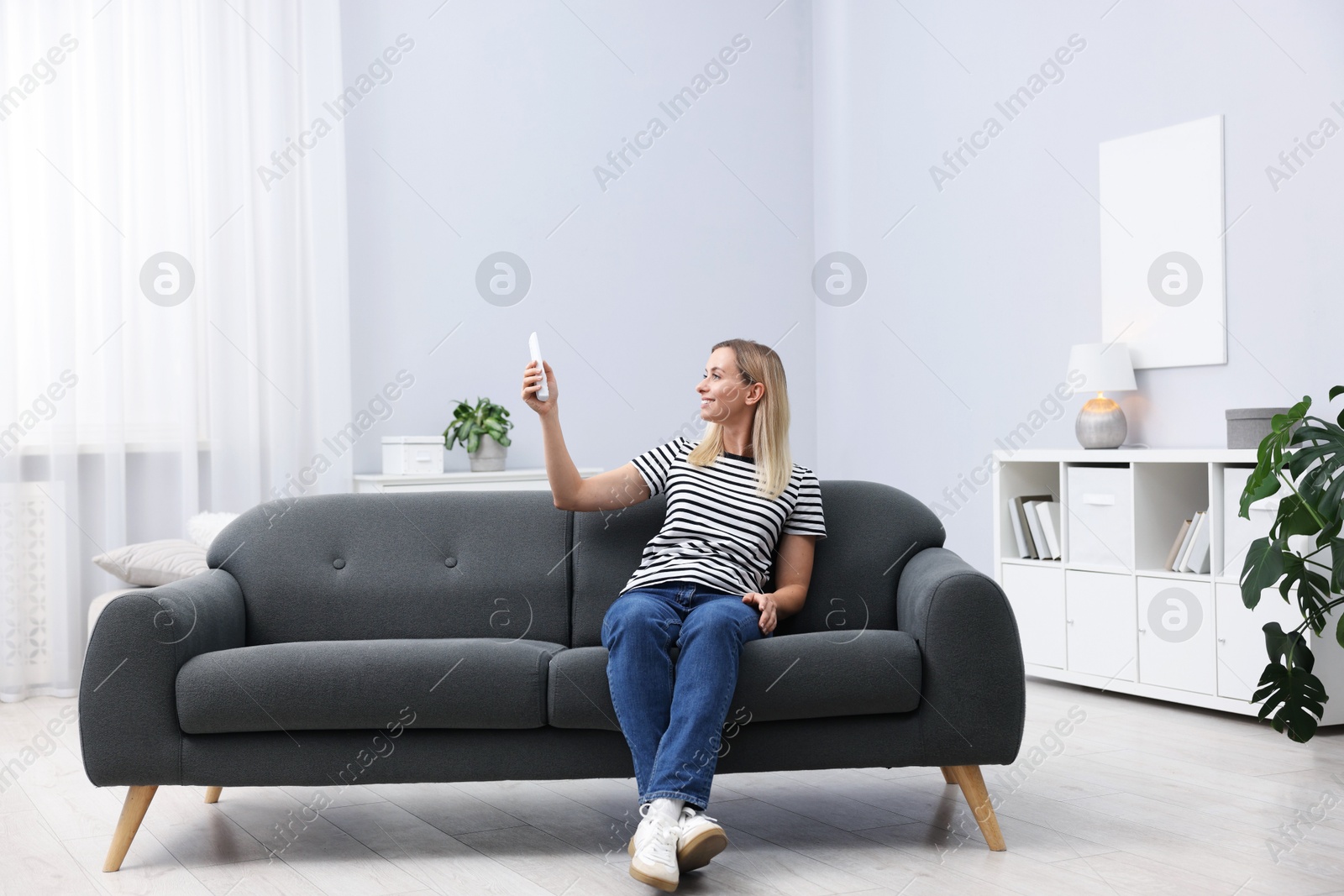 Photo of Woman using remote to control air conditioner on sofa at home