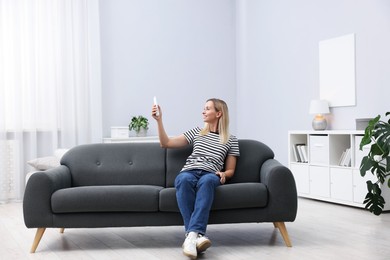 Photo of Woman using remote to control air conditioner on sofa at home