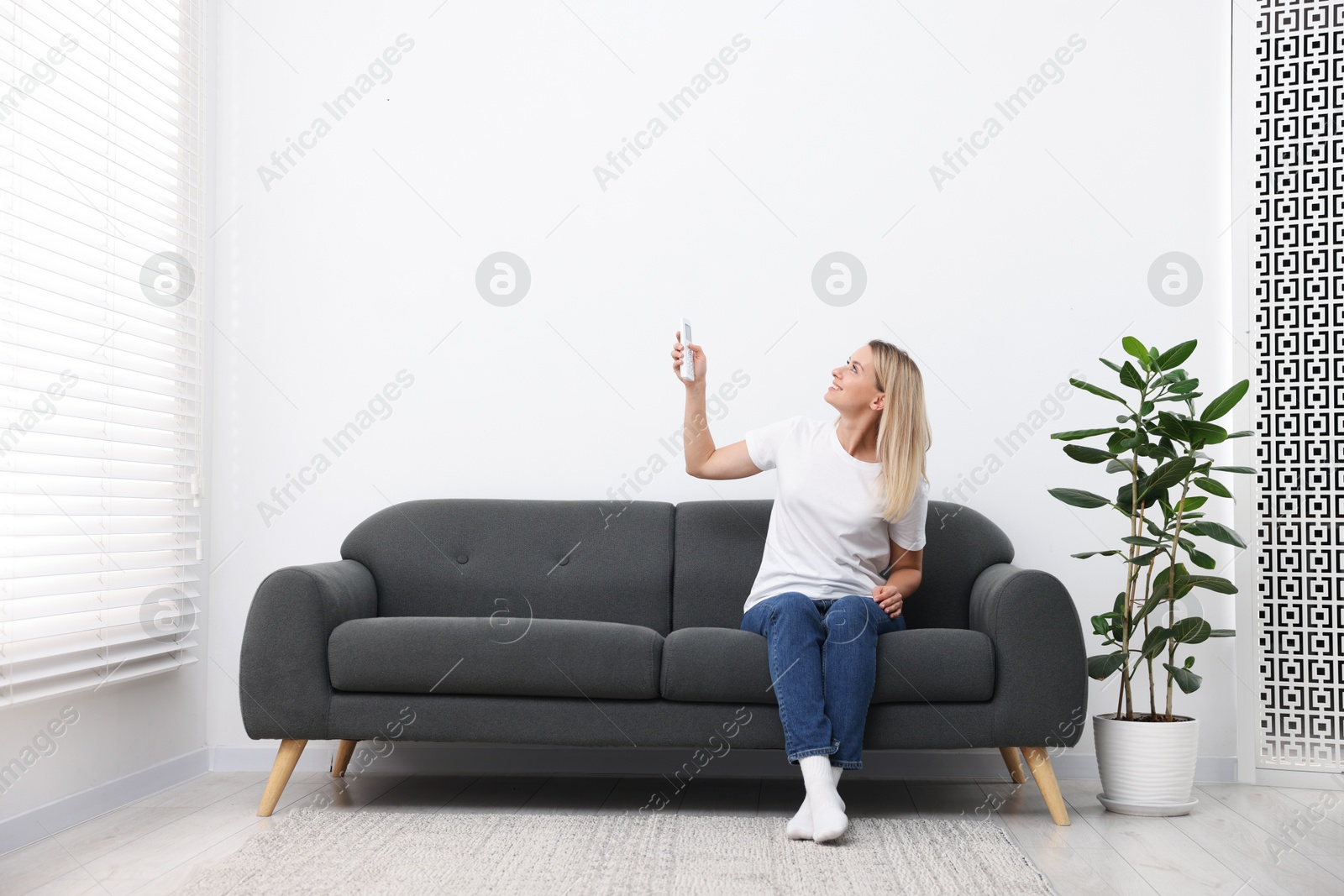 Photo of Woman using remote to control air conditioner on sofa at home. Space for text