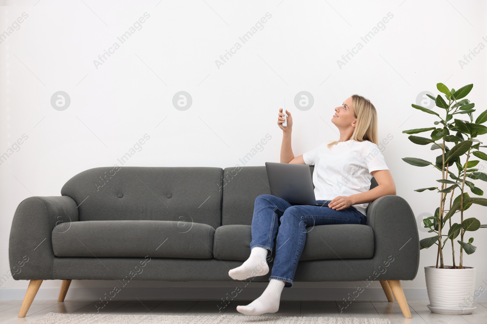 Woman with laptop using remote to control air conditioner on sofa at home. Space for text Photo of Woman with laptop using remote to control air conditioner on sofa at home. Space for text