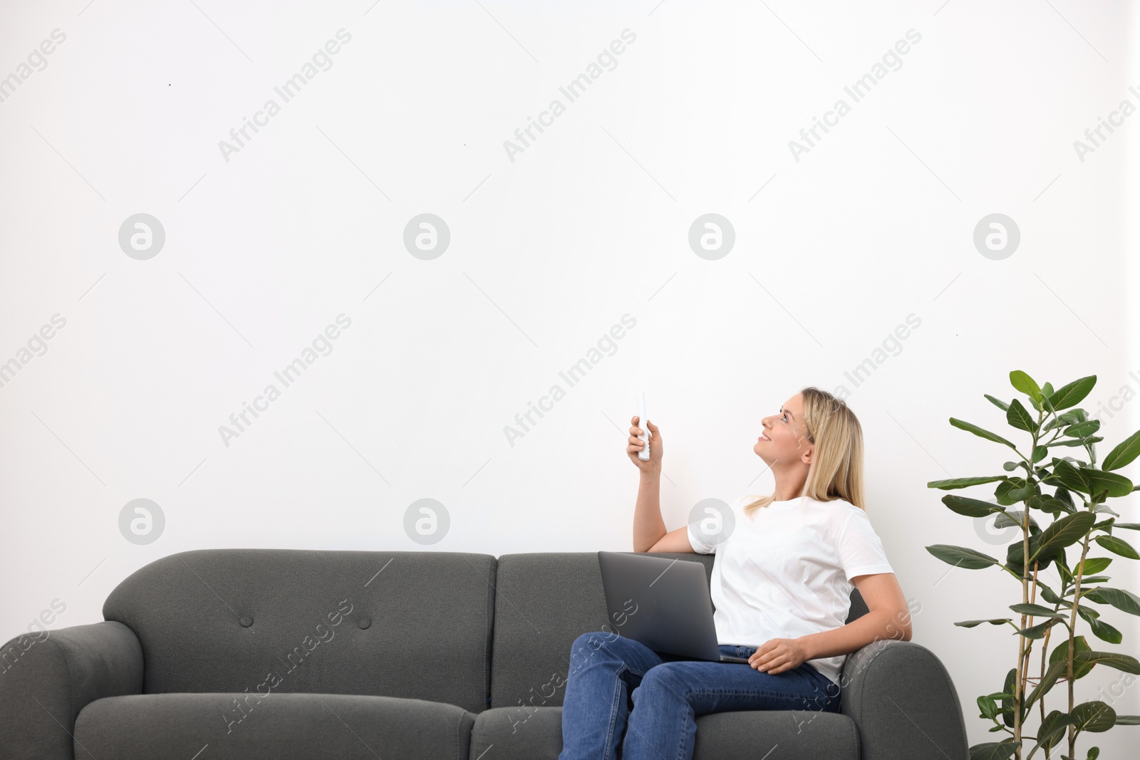 Woman with laptop using remote to control air conditioner on sofa at home. Space for text Photo of Woman with laptop using remote to control air conditioner on sofa at home. Space for text