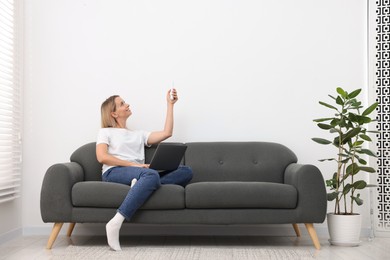 Photo of Woman with laptop using remote to control air conditioner on sofa at home