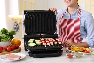 Photo of Woman cooking skewers with meat and vegetables on electric grill at home, closeup