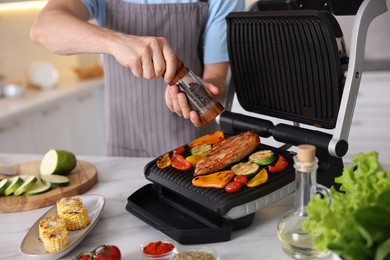 Photo of Man adding pepper to vegetables and meat cooking on electric grill in kitchen, closeup