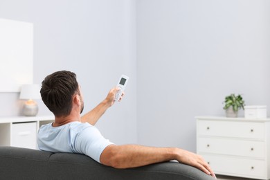 Photo of Man using remote to control air conditioner on sofa at home, back view. Space for text