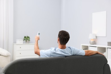 Photo of Man using remote to control air conditioner on sofa at home, back view