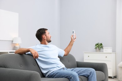 Photo of Man using remote to control air conditioner on sofa at home