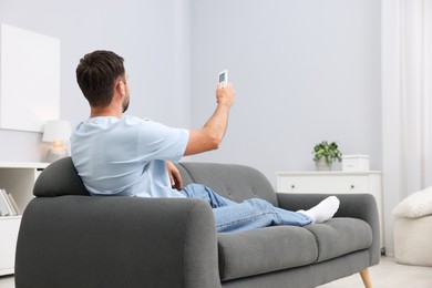 Photo of Man using remote to control air conditioner on sofa at home. Space for text
