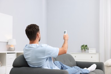 Photo of Man using remote to control air conditioner on sofa at home. Space for text