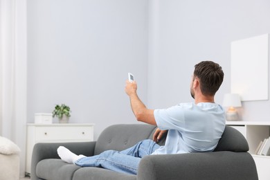 Photo of Man using remote to control air conditioner on sofa at home. Space for text