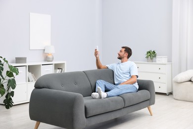 Photo of Man using remote to control air conditioner on sofa at home