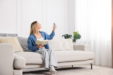 Photo of Woman with book using remote to control air conditioner on sofa at home