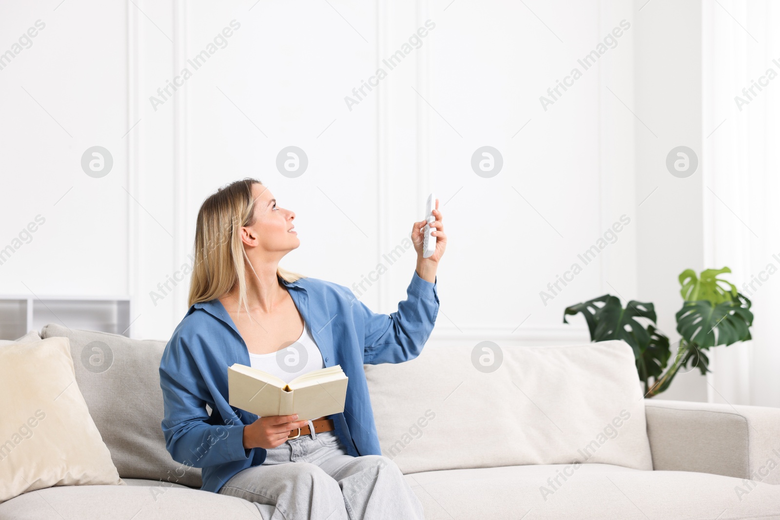 Photo of Woman with book using remote to control air conditioner on sofa at home
