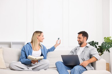 Photo of Couple with laptop and book using remote to control air conditioner on sofa at home