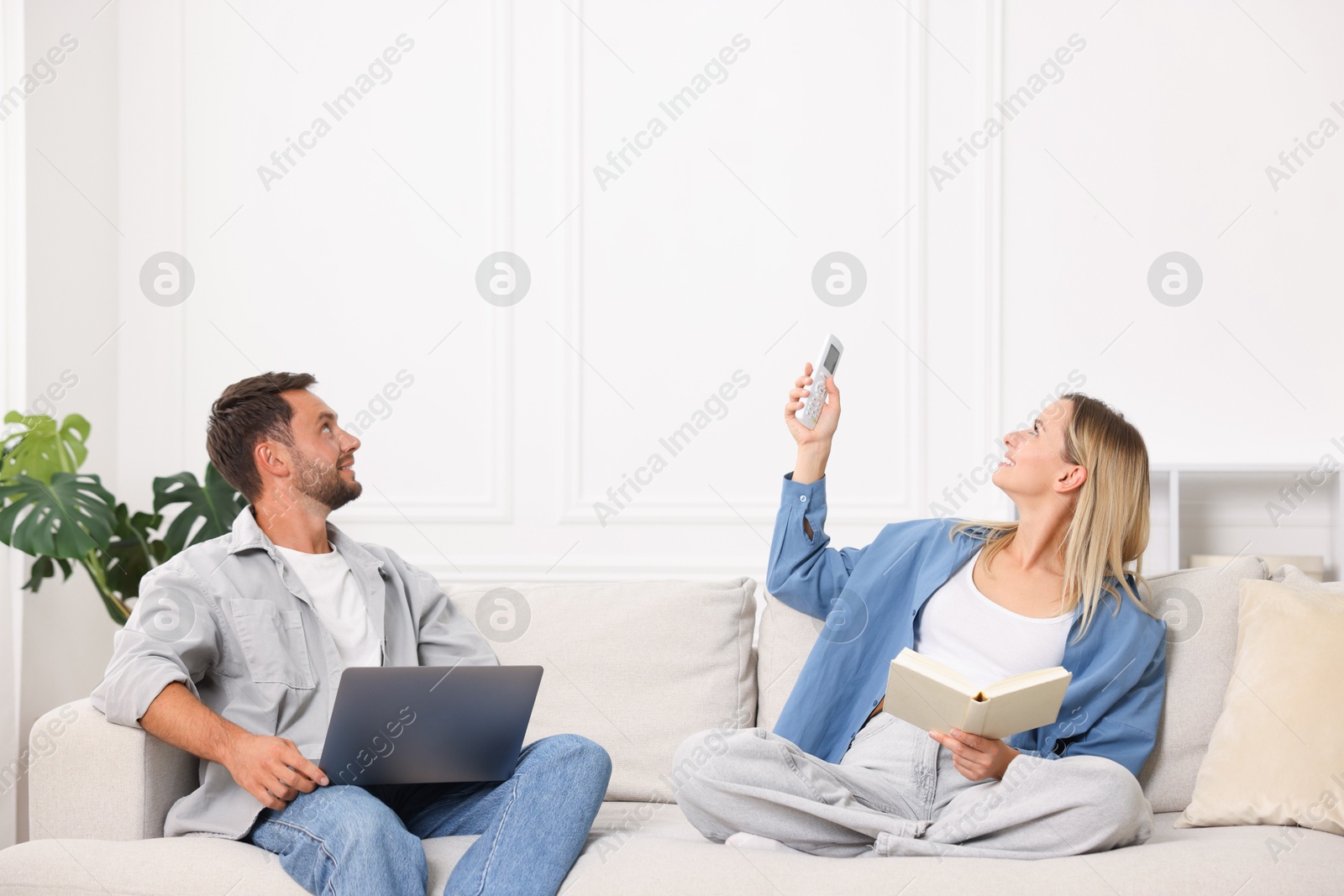 Photo of Couple with laptop and book using remote to control air conditioner on sofa at home