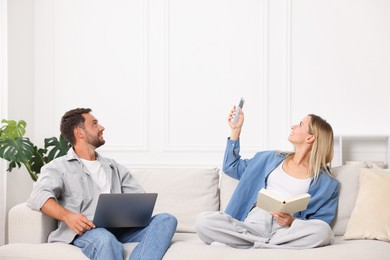 Photo of Couple with laptop and book using remote to control air conditioner on sofa at home