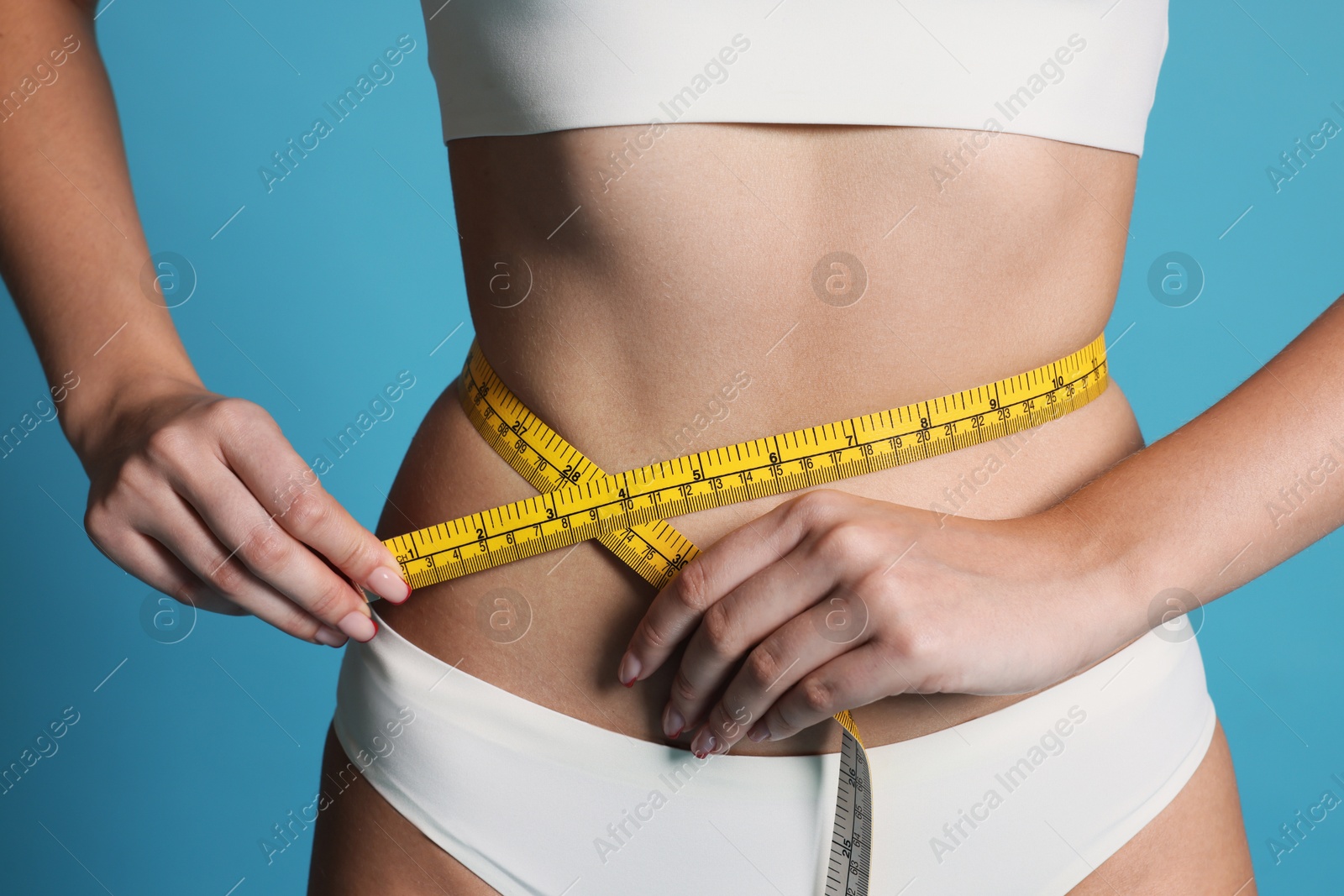 Photo of Young woman measuring her flat belly with tape on light blue background, closeup