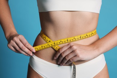 Photo of Young woman measuring her flat belly with tape on light blue background, closeup