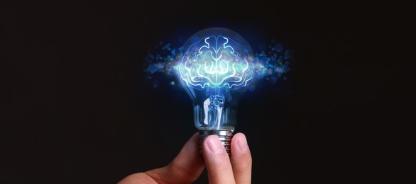Image of Man holding light bulb with brain illustration inside against black background, closeup. Idea concept